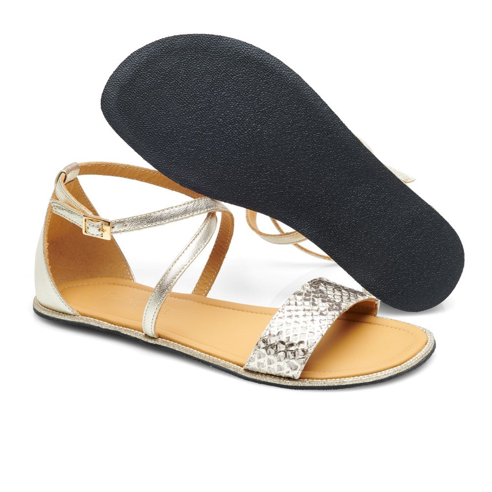 Die ZAQQ QEEP Gold Damensandalen bestechen durch ein elegantes Goldmetallic-Finish mit strukturierten Riemen und einer flachen schwarzen Sohle. Eine Sandale steht aufrecht, um das Riemchendesign hervorzuheben, während die andere umgedreht ist und die Sohle aus goldenem Nappaleder freigibt.