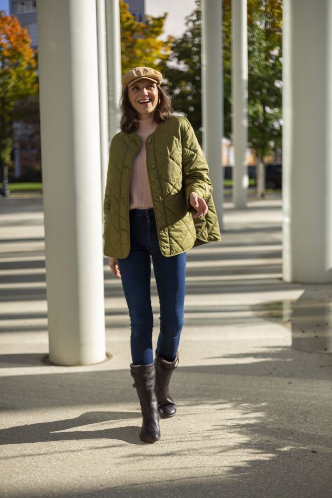 Eine Frau in einer grünen Steppjacke, Blue Jeans und ZAQQs ATTRAQT Winter Brown-Stiefeln geht im Freien spazieren. Sie lächelt und genießt den Komfort ihres Outfits, ergänzt durch eine beige Mütze. Hohe weiße Säulen und Bäume mit Herbstlaub unterstreichen den ruhigen Hintergrund.