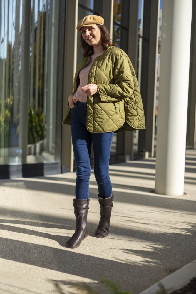 Eine Frau in einer grünen Steppjacke, Blue Jeans und ATTRAQT Winter Brown Boots von ZAQQ steht draußen im Sonnenlicht. Mit einer beigefarbenen Baskenmütze und einem Lächeln auf ihrem Gesicht strahlt sie Behaglichkeit aus. Glasfenster und Säulen bilden den Hintergrund für diese Szene aus Stil und Tragekomfort.