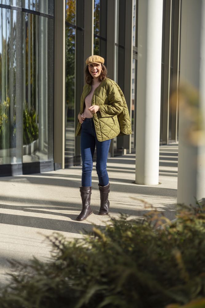 Eine Person in olivgrüner Steppjacke, beiger Baskenmütze und Jeans steht lächelnd im Freien neben hohen Fenstern und Säulen. Ihre ZAQQ ATTRAQT Winter Brown-Stiefel bieten außergewöhnlichen Tragekomfort, während Schatten auf dem Bürgersteig tanzen. Im Vordergrund sind Büsche zu sehen.