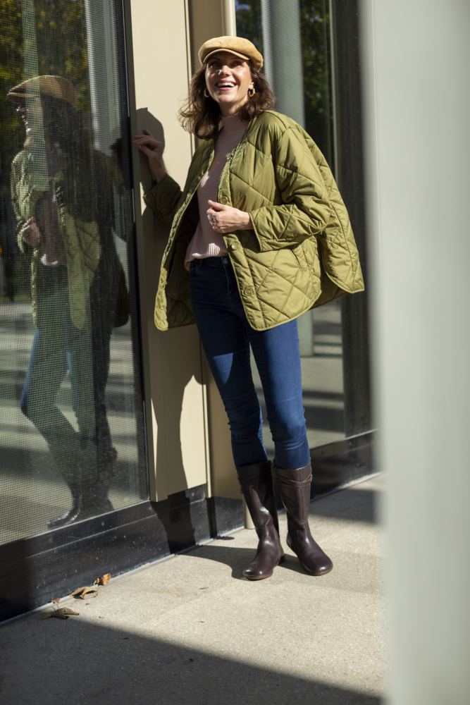 Eine Frau in einer hellgrünen Steppjacke, Bluejeans und braunen ATTRAQT Nappa Boots von ZAQQ steht lächelnd neben einer Glaswand, in der sich ihr Bild spiegelt. Mit ihrer beigen Mütze scheint sie einen sonnigen Tag im Freien zu genießen.