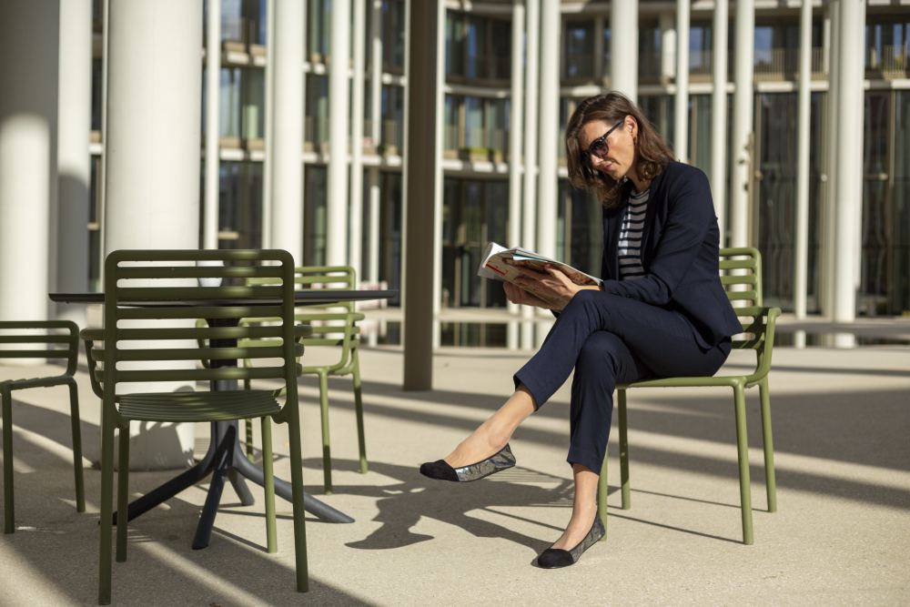 Eine Frau in einem marineblauen Anzug und mit Sonnenbrille sitzt auf einem grünen Gartenstuhl und liest ein Buch. Ihre schwarzen ZAQQ TWIST Derby-Schuhe ruhen bequem auf dem Boden, während sie mit moderner Architektur und Glasfenstern im Hintergrund das Sonnenlicht genießt.