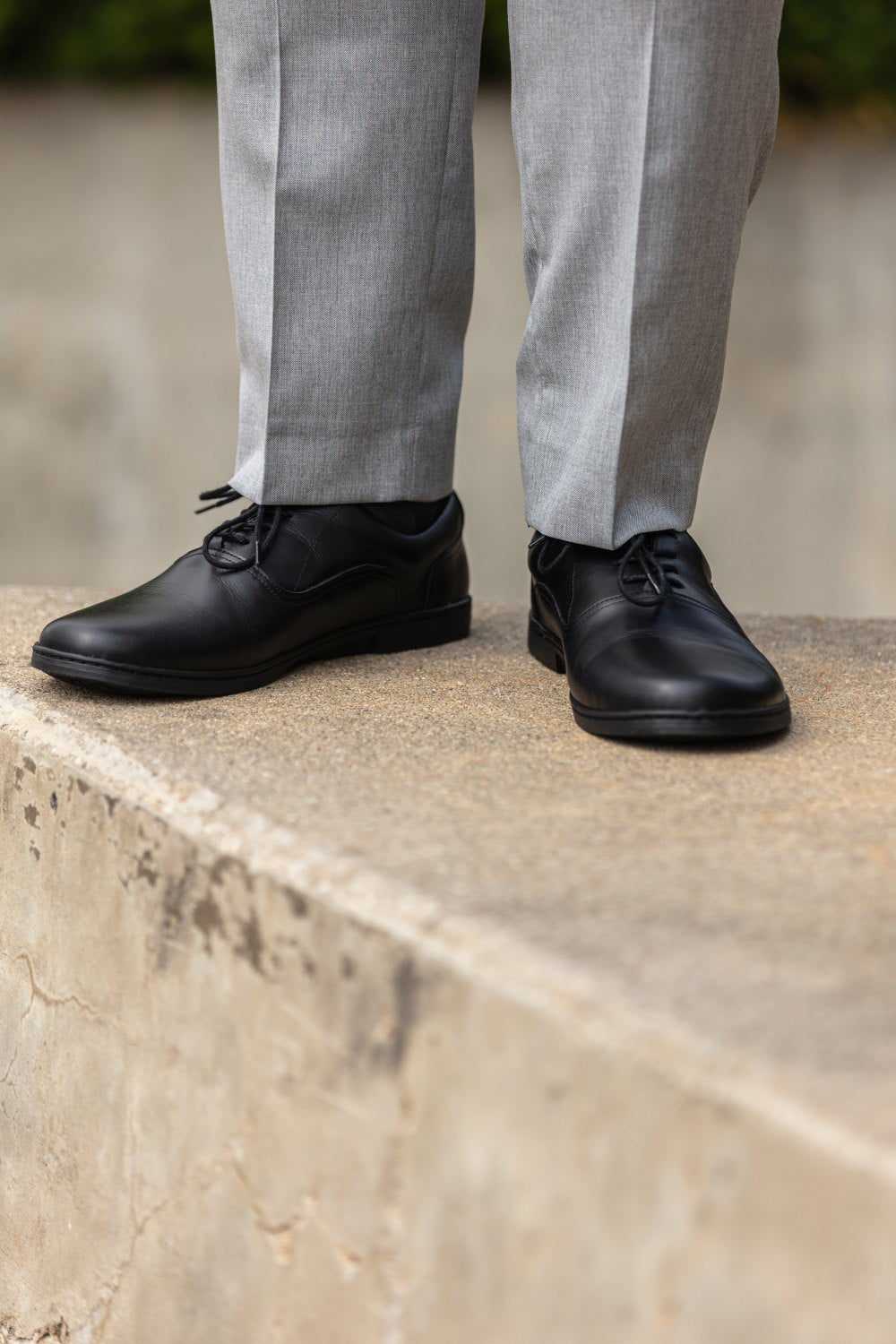 Eine Person in grauer Hose und schicken APEQ Black Schuhen von ZAQQ aus poliertem Leder steht auf einer Betonkante. Der Fokus liegt auf der unteren Hälfte, die das elegante Schuhwerk in Szene setzt. Der unscharfe Hintergrund hebt die Schuhe und einen Teil der Hose raffiniert hervor.