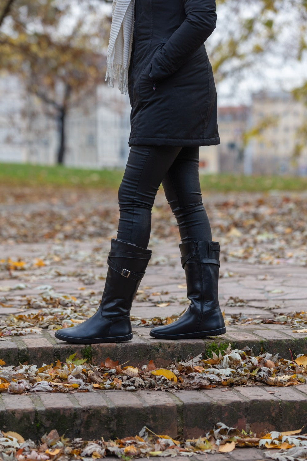 Eine Person in schwarzen ATTRAQT Winter Black-Stiefeln von ZAQQ und einem passenden Mantel schlendert an einem Herbsttag auf einem mit Blättern bedeckten Kopfsteinpflasterweg in einem Park entlang. Gefallene Blätter liegen verstreut auf dem Boden, im Hintergrund sind Bäume mit spärlichem Laub zu sehen.