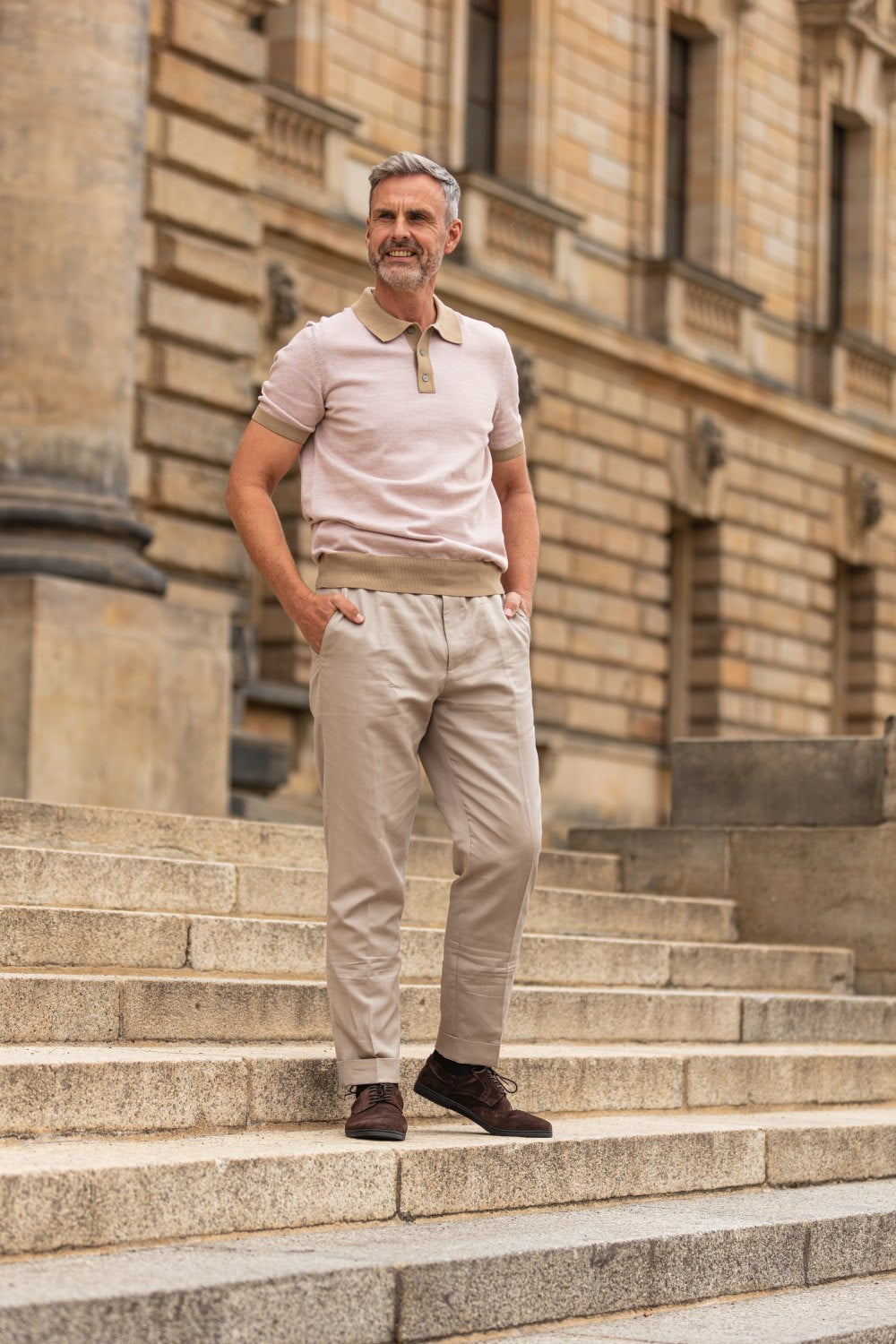 Ein Mann mit grauem Haar und Bart steht selbstbewusst auf Steinstufen vor einem historischen Gebäude. Er trägt ein beiges Poloshirt, eine passende Hose und die Schuhe BRIQ Brogue Velours Brown von ZAQQ, die Hände in den Taschen.