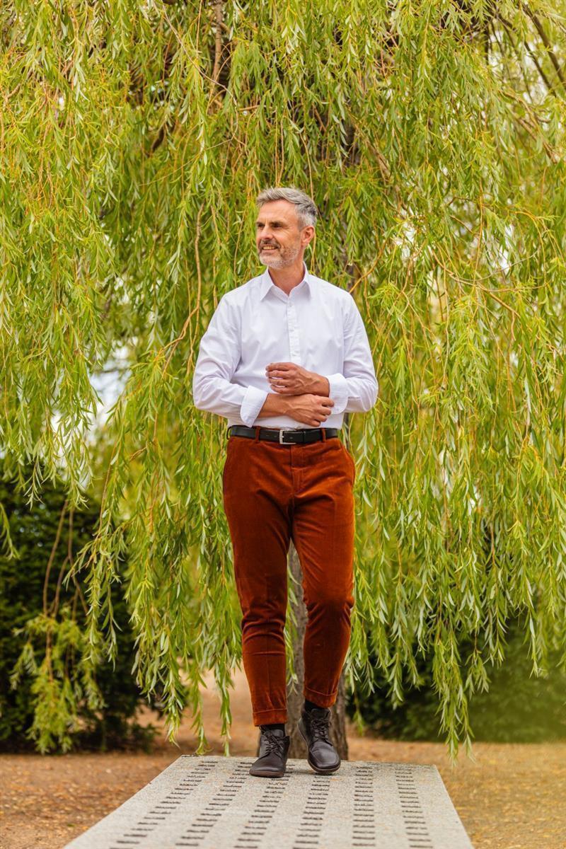 Ein Mann mit grauem Haar und Bart steht draußen auf einer gemusterten Plattform. Er trägt die Schuhe NEWQUAY Brown von ZAQQ, ein weißes Hemd und braune Hosen. Er wird von herabhängenden Weidenzweigen in einer ruhigen, natürlichen Umgebung eingerahmt.