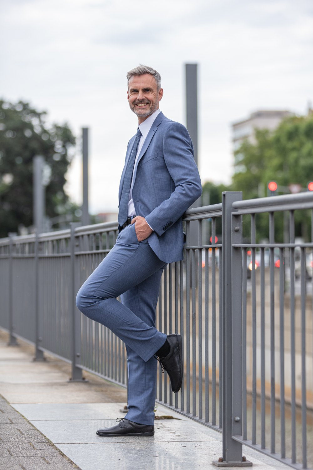 Ein Mann im blauen Anzug lehnt selbstbewusst an einem Metallgeländer auf einem Bürgersteig in der Stadt. Seine schwarzen ZAQQ PEAQ Brogue-Schuhe verleihen ihm eine elegante Note. Er lächelt, während er mit gekreuzten Beinen posiert. Im Hintergrund sind verschwommene Bäume, Gebäude und dezenter Stadtverkehr zu sehen.