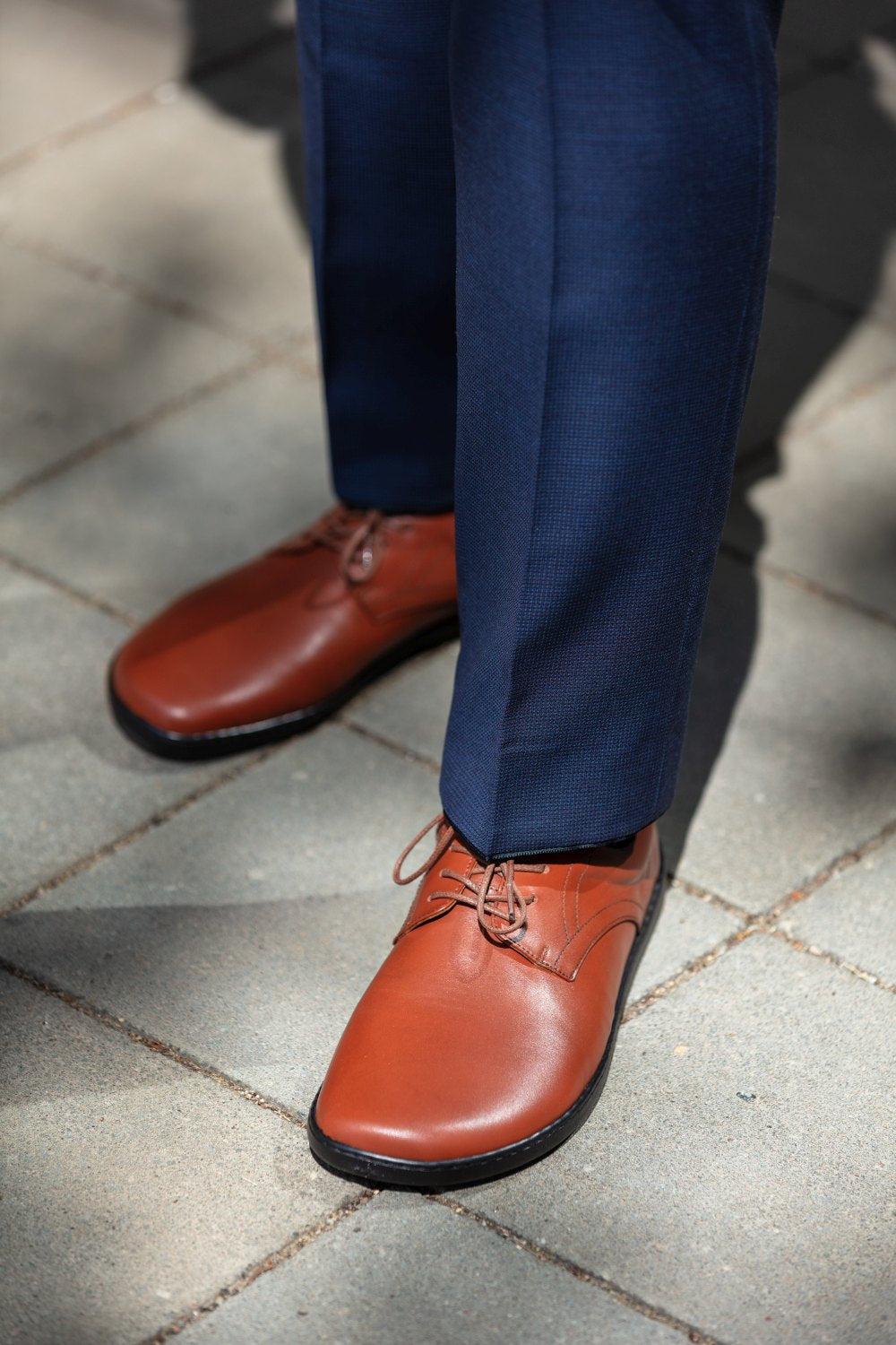 Eine Person in eleganten ZAQQ PEAQ Cognac-Lederschuhen und dunkelblauen Hosen steht auf einem gefliesten Gehweg, die Beleuchtung wirft weiche Schatten.