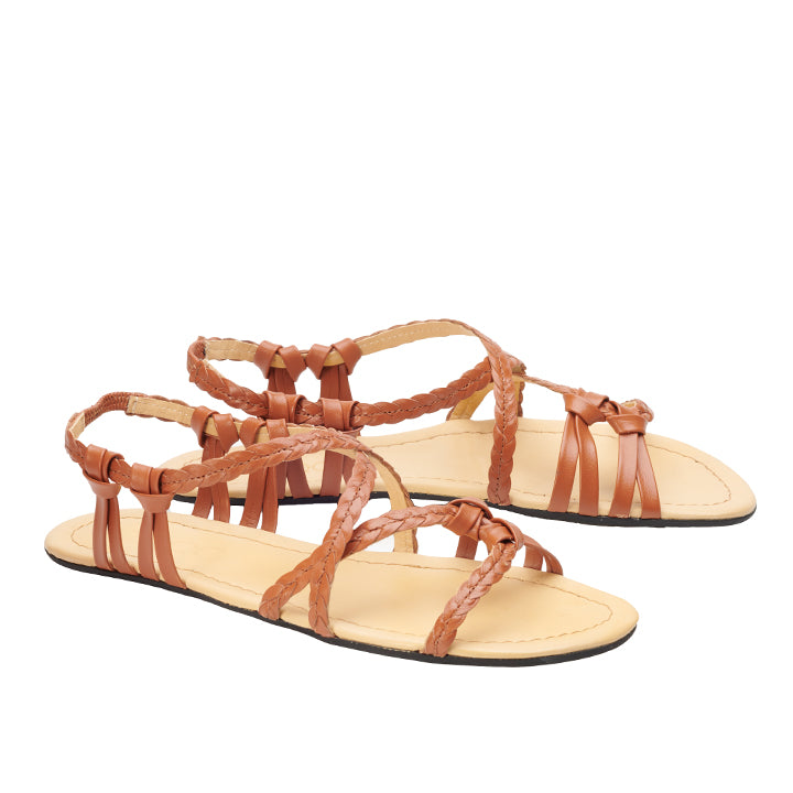 Das Modell QALMA Cognac von ZAQQ ist ein Paar braune Riemchensandalen aus hochwertigem Nappaleder mit geflochtenen und flachen Riemen, einer hellbraunen Innensohle für außergewöhnlichen Komfort und einer flachen schwarzen Sohle, alles auf weißem Hintergrund.