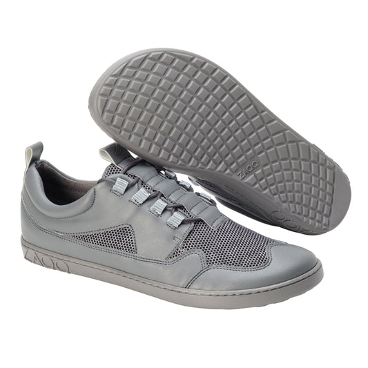 Ein Paar QARLY Grey-Sneaker von ZAQQ, bei denen ein Schuh aufrecht steht und der andere auf der Seite liegt, sodass die strukturierte Sohle sichtbar wird. Das Design umfasst einen Mesh-Abschnitt, Schnürsenkeldetails und ein Branding an der Seite und der Sohle. Diese Schuhe sind aus veganem Kunstleder gefertigt und verbinden nahtlos Stil mit Nachhaltigkeit.