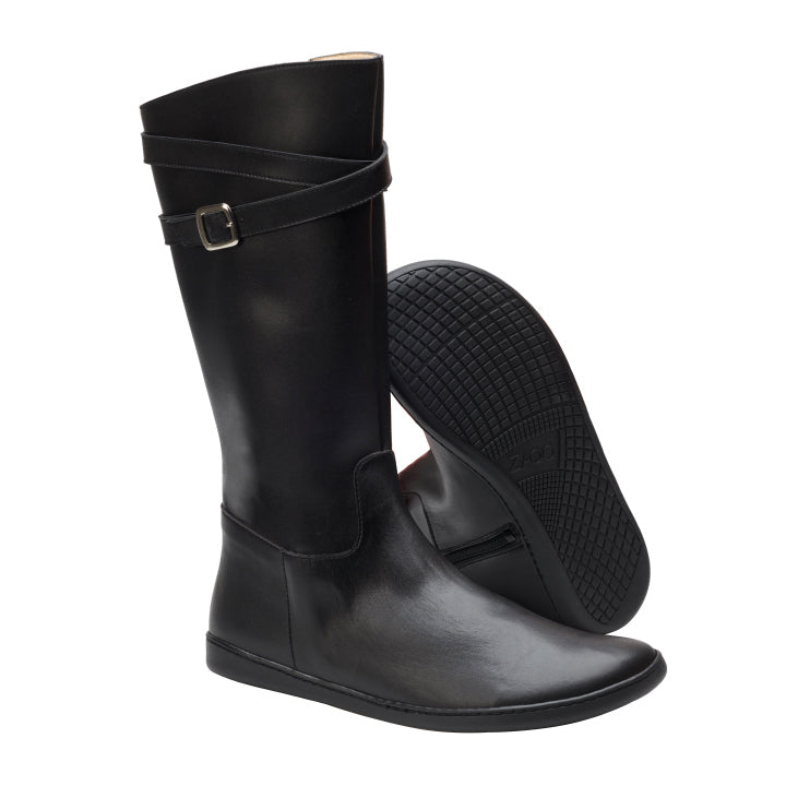 Das Modell ATTRAQT Black ist ein Paar veganer kniehoher Stiefel von ZAQQ und zeichnet sich durch ein minimalistisches Design mit Riemen und Schnalle an der Oberseite aus. Ein Stiefel steht aufrecht, während der andere angewinkelt ist, wodurch die strukturierte Sohle hervorgehoben wird.