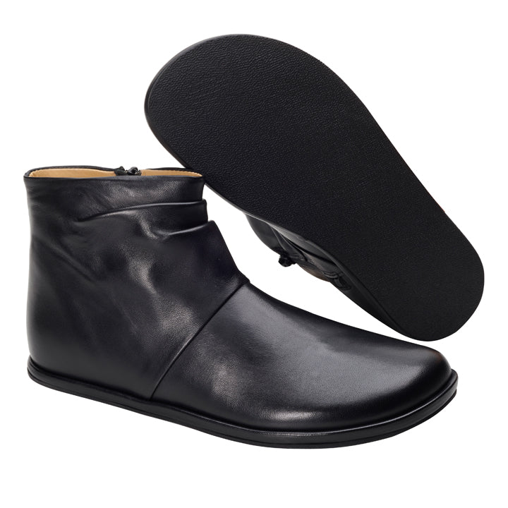 Ein Paar elegante schwarze Stiefeletten von ZAQQ QOURTNEY. Ein Stiefel steht aufrecht und betont die glatte Lederaußenseite, während der andere mit der Sohle nach oben gezeigt wird, um die strukturierte schwarze Sohle hervorzuheben. Das Design zeichnet sich durch eine moderne und minimalistische Ästhetik aus.