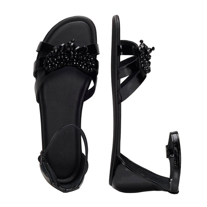 Die ZAQQ QISELLE Black Sandalen bestechen durch glänzende Riemchen, die mit Perlenverzierungen verziert sind. Eine Sandale, die einer Barfuß-Sandale ähnelt, ist von oben zu sehen, die andere von der Seite. Sie bestechen durch elegante Fesselriemchen und eine bequeme flache Sohle.