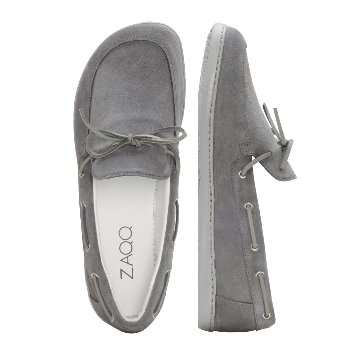 Die Loafer QEA Velours Grey von ZAQQ sind aus hochwertigem grauem Wildleder gefertigt und haben oben ein elegantes Schleifendetail. Ein Schuh ist von oben zu sehen und zeigt die Handwerkskunst, während der andere von der Seite zu sehen ist. Auf der Innensohle ist der Markenname ZAQQ deutlich zu erkennen.