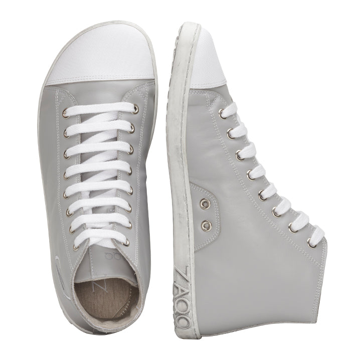 Ein Paar stylische CHUQQS Grey High-Top-Sneaker von ZAQQ mit Obermaterial aus hellgrauem Nappaleder und weißen Gummisohlen. Ein Schuh ist von der Seite zu sehen und zeigt das ZAQQ-Logo in der Nähe der Sohle, während der andere Schuh von oben zu sehen ist und seine weißen Schnürsenkel betont.