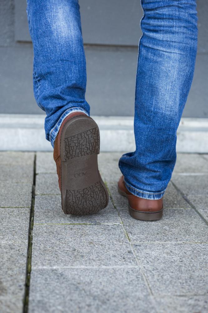 Eine Person in Blue Jeans und BRIQ Brogue Antique Cognac Schuhen von ZAQQ läuft auf einem gefliesten Bürgersteig, wobei der Fokus auf den Unterschenkeln und den Schuhsohlen liegt.