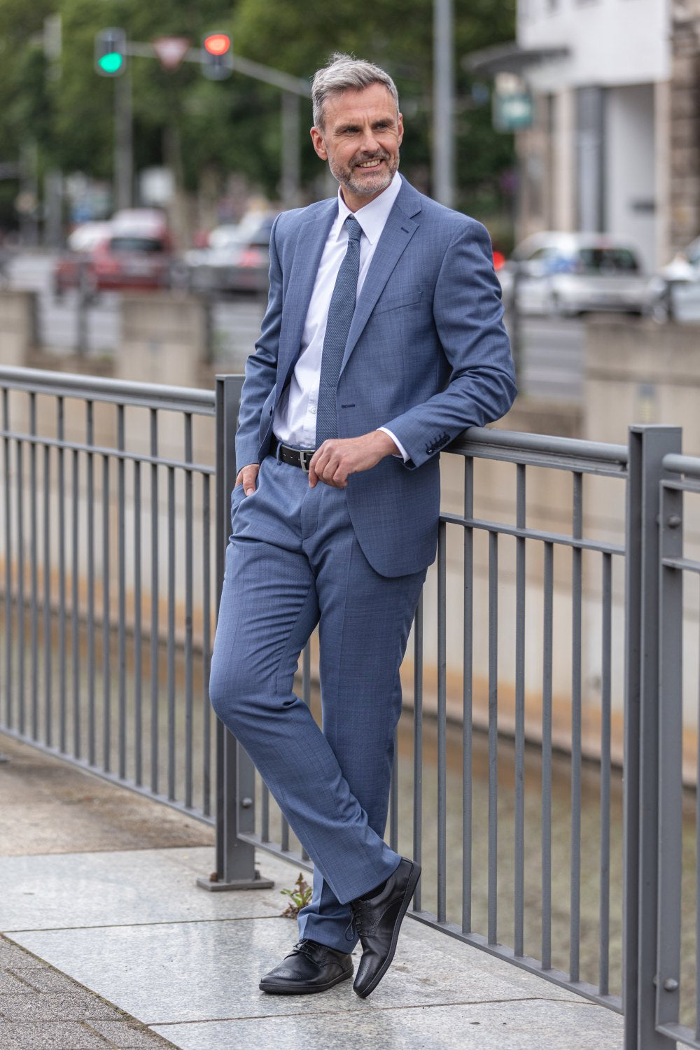 Ein Mann in blauem Anzug und Krawatte steht lässig an einem Geländer auf einem Bürgersteig in der Stadt, mühelos stilvoll in seinen schwarzen ZAQQ PEAQ Brogue-Schuhen. Er lächelt und hat eine Hand in der Tasche, während Autos und Bäume einen verschwommenen Hintergrund bilden.