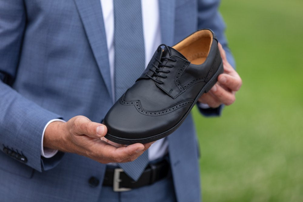 Eine Person im blauen Anzug hält den eleganten Schuh BRIQ Brogue Black von ZAQQ und präsentiert sein klassisches schwarzes Lederdesign mit Brogue-Details vor einem verschwommenen grünen Hintergrund.