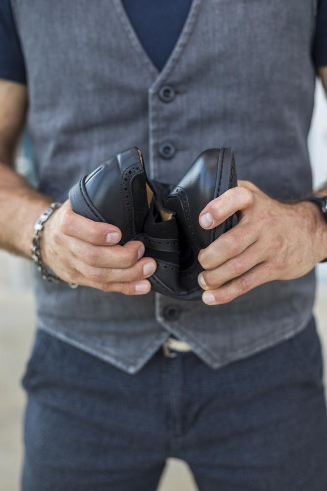 Eine Person in grauer Weste und blauem Hemd hält ein Paar BRIQ Brogue Deep Black Schuhe von ZAQQ, wobei die Absätze zusammen in den Händen positioniert sind. Der unscharfe Hintergrund lenkt die Aufmerksamkeit auf die Schuhe und die Kleidung der Person.