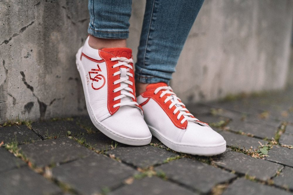 Eine Person trägt die CEDRIQ Red Sneaker von ZAQQ mit roten Akzenten und Schnürsenkeln und steht auf einem Kopfsteinpflasterweg. Die Sneaker zeigen markante Buchstaben und Symbole, die für den einzigartigen Stil von ZAQQ charakteristisch sind. Kombiniert mit Blue Jeans bieten diese Barfuß-Sneaker dank ihrer flexiblen Kautschuksohle Komfort.