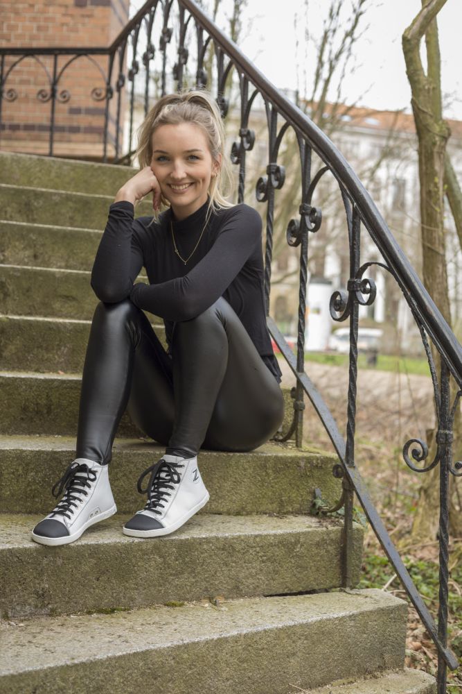 Eine Frau mit blondem Haar, einem eleganten schwarzen Outfit und stylischen weißen CHUQQS-Sneakern von ZAQQ, sitzt auf einer Außentreppe, lächelt und legt ihre Hand unter ihr Kinn. Im Hintergrund sind Bäume und ein Gebäude zu sehen.