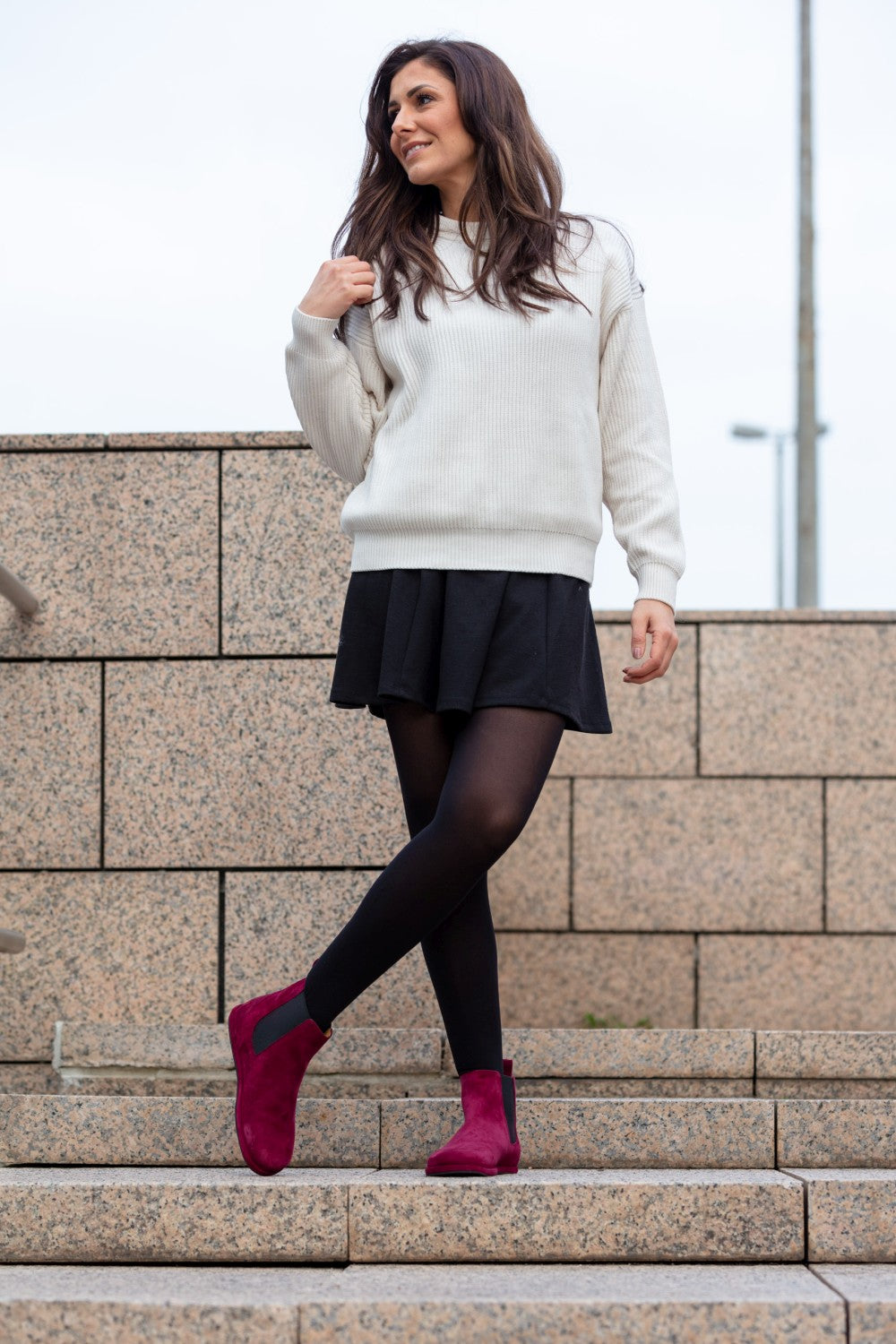 Eine Person mit langen dunklen Haaren steht selbstbewusst auf Steinstufen und trägt einen beigen Pullover, einen schwarzen Rock und ZAQQ EQUITY Berry-Stiefel. Schwarze Strumpfhosen ergänzen ihr burgunderfarbenes Schuhwerk vor dem Hintergrund von Steinmauern und einem bewölkten Himmel.