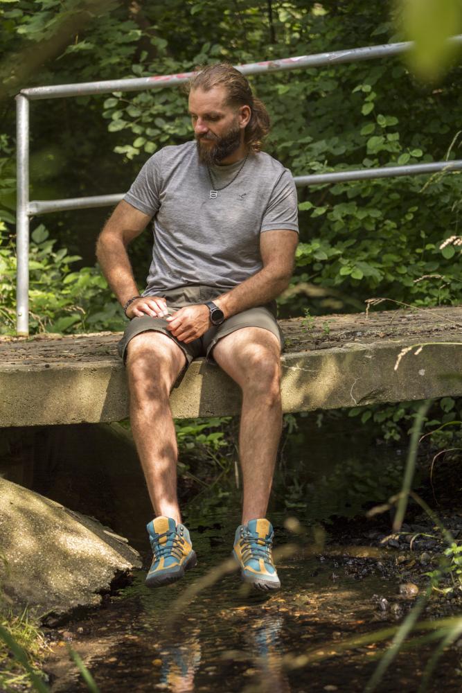 Ein Mann mit langen Haaren und Bart sitzt auf einer Betonbrücke in einem Waldgebiet. Er trägt ein graues T-Shirt, Shorts und bunte ZAQQ ESQAPE Blue Orange Waterproof-Sneaker und blickt auf einen kleinen Bach unter ihm. Dichtes grünes Laub umgibt die ruhige Szene.