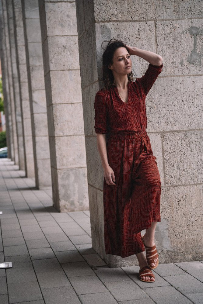 Eine Frau in einem langen, strukturierten roten Kleid lehnt an einer Steinmauer in einer Kolonnade, ihre Hand ruht auf ihrem Kopf. Sie steht auf einem gepflasterten Gehweg, trägt die minimalistischen FLAQE Brown Sandalen von ZAQQ aus Nappaleder und blickt nachdenklich in die Ferne.