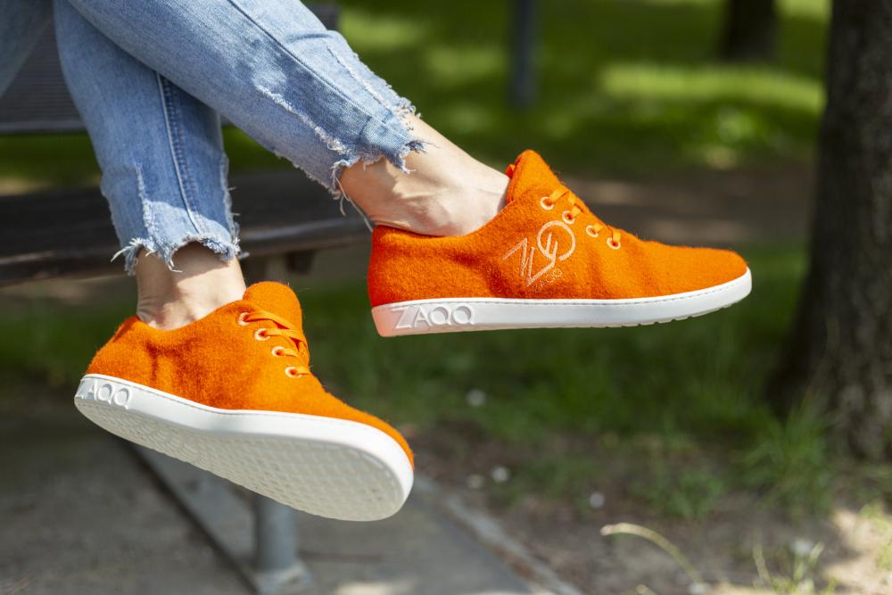 Eine Person trägt LIQE Orange-Sneaker von ZAQQ, in leuchtendem Orange mit weißen Sohlen und Branding, sitzt auf einer Bank und genießt das Barfußgefühl. Die Sneaker sind mit ausgefransten Bluejeans kombiniert, vor einem Hintergrund aus grünem Gras und Bäumen, der an eine Parkkulisse erinnert.