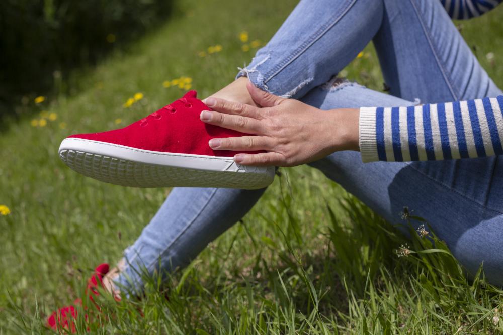 Eine Person sitzt im Gras. Sie trägt blaue Jeans und einen gestreiften Pullover und hält sich den Knöchel. Ihre roten Sneaker sind die LIQE Race von ZAQQ, mit einer bequemen Barfußsohle. Im Hintergrund sind Löwenzahn und grünes Gras zu sehen.