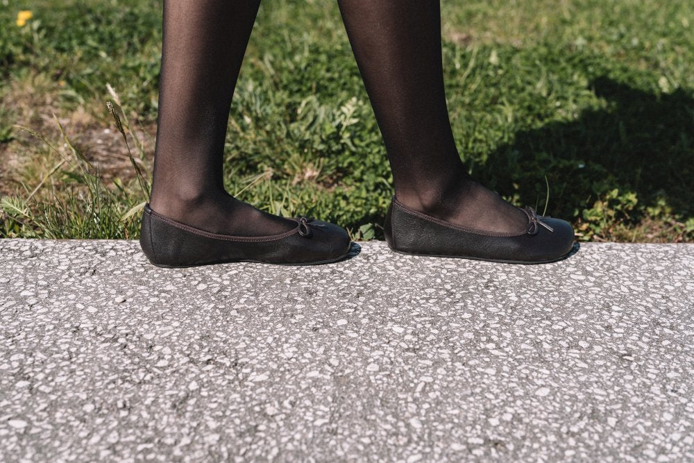 Eine Person in schwarzen Strumpfhosen und eleganten dunkelbraunen LOOQ-Ballerinas von ZAQQ schreitet selbstbewusst über einen strukturierten Betonweg neben einer Rasenfläche. Der Fokus liegt weiterhin auf ihren anmutigen Beinen und selbstbewussten Schritten.