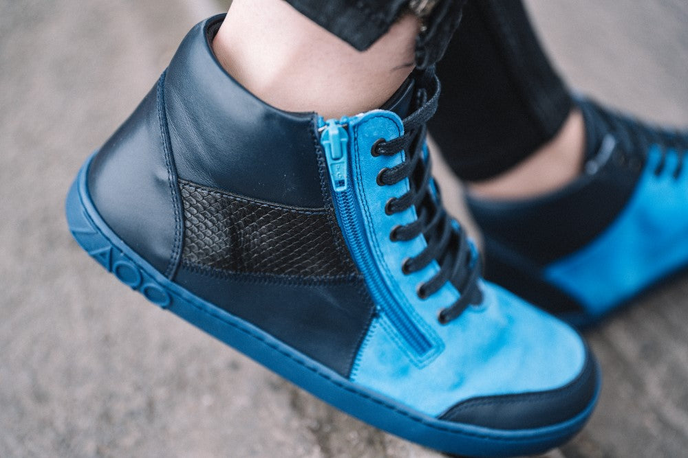 Eine Nahaufnahme einer Person, die blaue und schwarze LUQQ Azur High-Top-Sneaker von ZAQQ trägt, zeigt ein stilvolles Design mit Reißverschluss und strukturiertem Einsatz an der Seite. Mit leuchtend blauen Akzenten fangen diese eleganten Sneaker die Essenz des Halber-Sneaker-Designs ein und eignen sich perfekt zum Gehen auf einer Betonoberfläche.