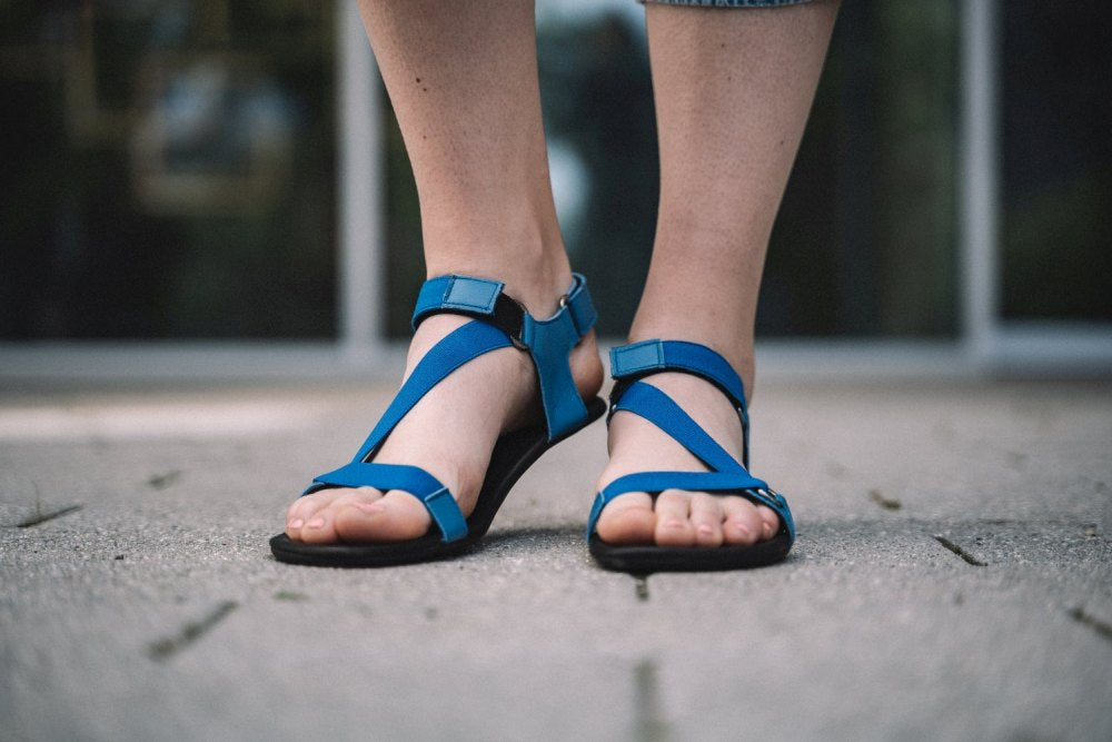 Eine Person in blauen ZAQQ MIQA Sandalen mit bequemem Klettverschluss steht auf einem strukturierten Bürgersteig. Ihre Unterschenkel und Füße sind vor dem Hintergrund eines verschwommenen Gebäudes mit Andeutungen von Fensterreflexionen zu sehen.