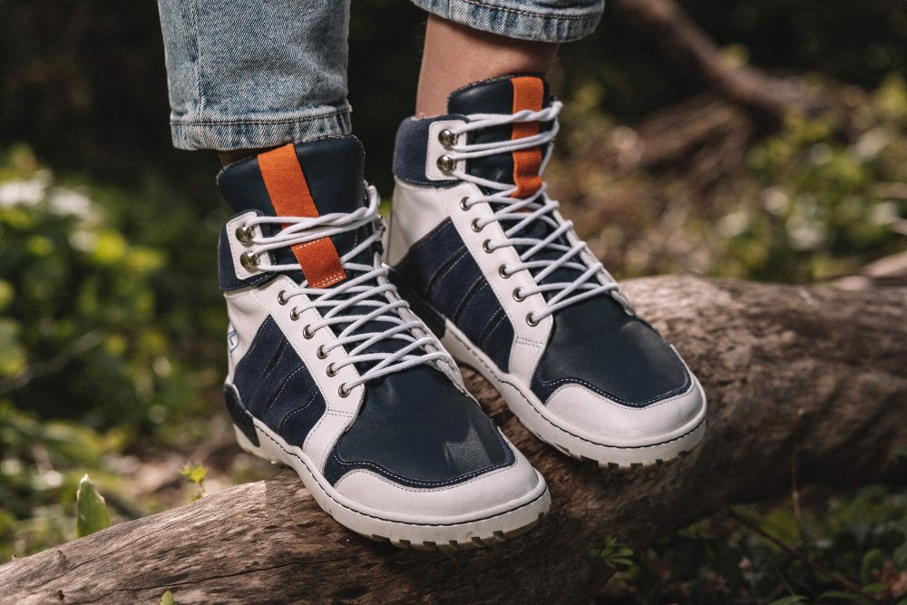 Ein Paar wasserdichte MONTBLANQ Blue White High-Top-Sneaker von ZAQQ aus Nappaleder mit blauen, weißen und orangefarbenen Akzenten schmücken die Füße einer Person, die selbstbewusst auf einem großen umgestürzten Ast steht. Die hochgekrempelten Jeans ergänzen das leuchtend grüne Laub im Hintergrund perfekt und machen diese Sneaker zu einem idealen Outdoor-Schuh für Abenteuer.