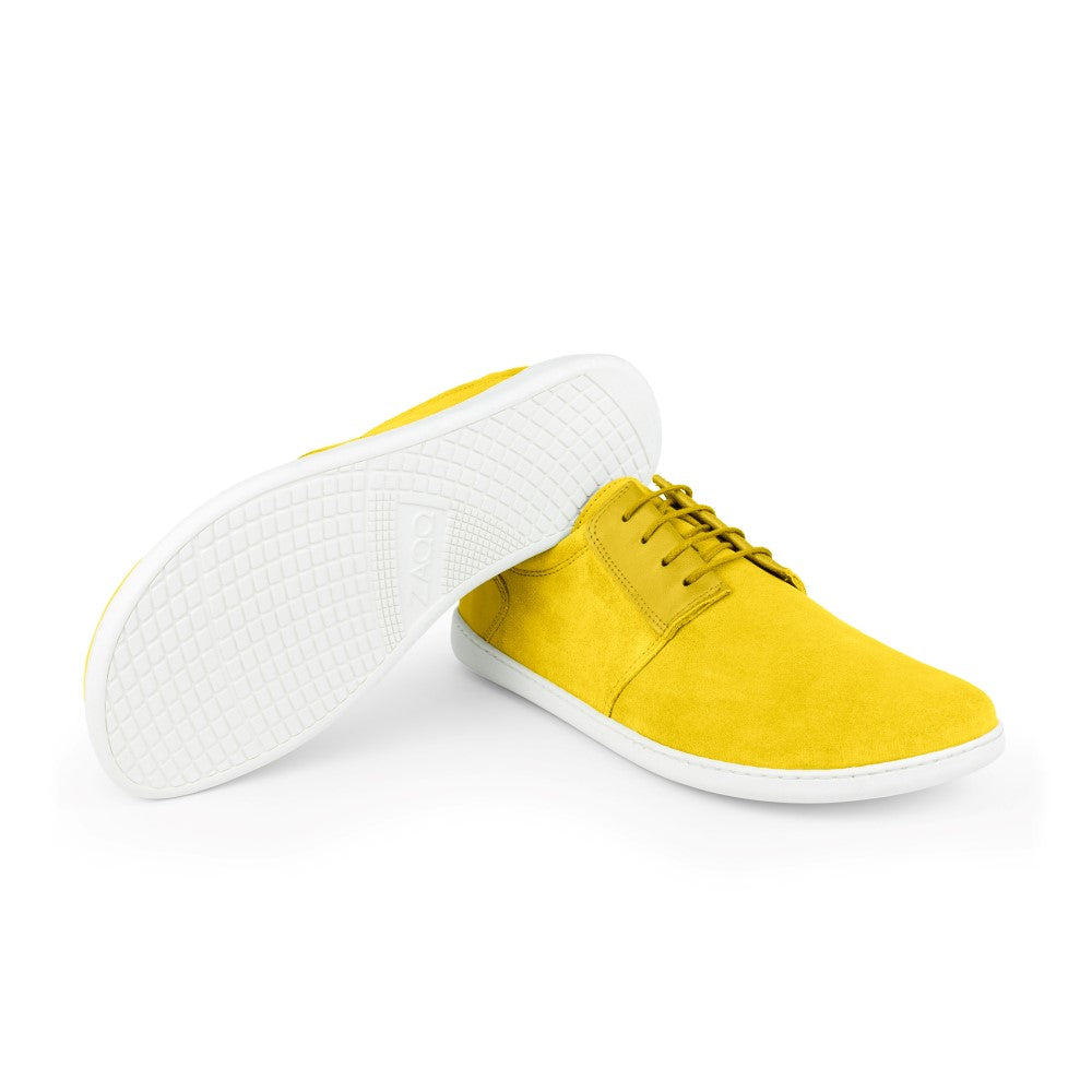 Wir präsentieren die PIQUANT Yellow Sneaker von ZAQQ in einem auffälligen Zitronengelb mit kontrastierenden weißen Sohlen. Ein Schuh steht aufrecht, während der andere auf der Seite liegt und eine markante Sohle mit Gittermuster zeigt. Diese Schnürsneaker aus hochwertigem Veloursleder strahlen ein elegantes und modernes Design aus, ideal für alle, die Wert auf Barfußkomfort legen.