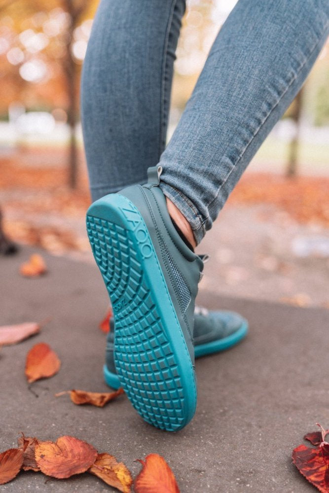 Eine Person steht auf einem mit verstreuten roten und orangen Herbstblättern bedeckten Weg und trägt blaue ZAQQ Barfuß-Sneaker mit QARLY Turquoise Sohlen. Der Fokus liegt auf der Schuhsohle, während die Bäume und das Gras im Hintergrund kunstvoll verschwimmen.