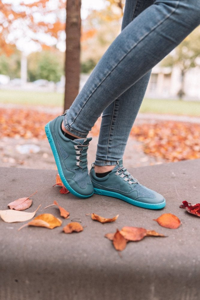 Eine Person, die ZAQQs QARLY Turquoise-Sneaker und Jeans trägt, steht auf einem Betonvorsprung, umgeben von herbstlichen Blättern. Im Hintergrund sind ein Baum und eine Grasfläche zu sehen, die das Wesen der Jahreszeit perfekt einfangen.