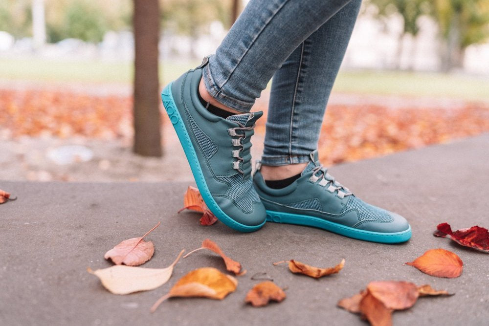 Eine Person in türkisfarbenen ZAQQ QARLY-Sneakers steht auf einem gepflasterten Weg, umgeben von verstreutem Herbstlaub. Ihre Beine in Blue Jeans ergänzen die Sneakers, während eine verschwommene Parkkulisse der Szene Tiefe verleiht.