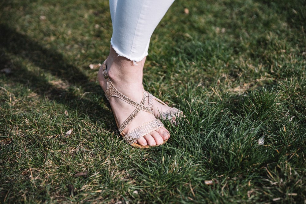 Eine Person in einer weißen Hose und den QEE-Taupe-Sandalen von ZAQQ, die ein modernes Design und eine flexible Sohle aufweisen, steht auf grünem Gras.