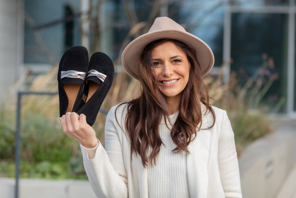 Eine Frau mit beigem Hut und weißem Pullover lächelt und hält ein Paar schwarze QERP Velours-Loafer von ZAQQ hoch, die mit eleganten Schleifen versehen und aus glattem Veloursleder gefertigt sind. Sie steht im Freien vor einem unscharfen Hintergrund mit Pflanzen und einem Gebäude.