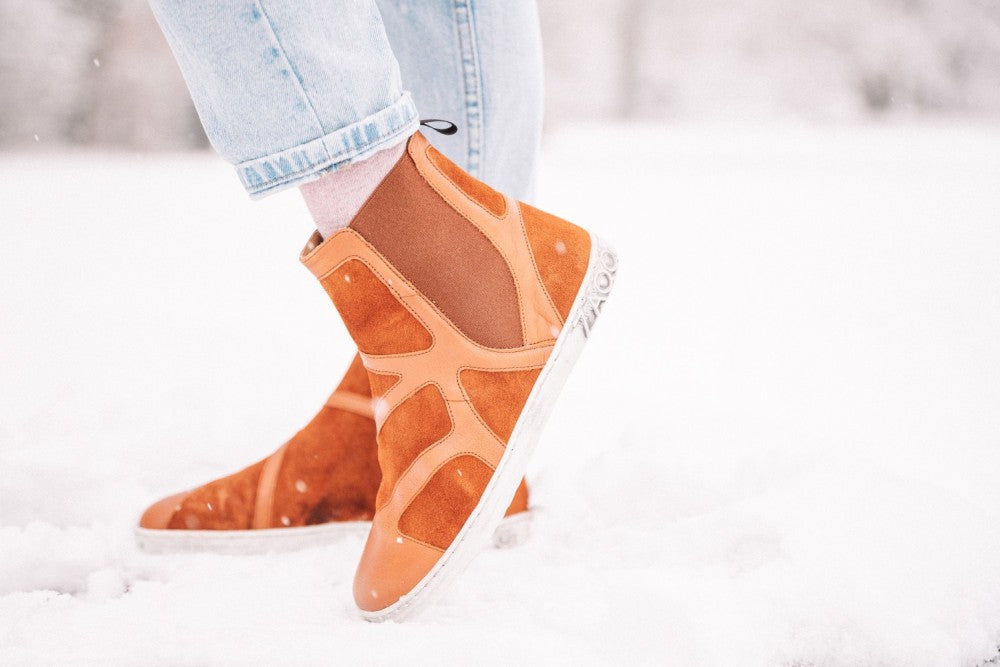 Eine Person trägt die stylischen QEY WEST Cognac Sneakers von ZAQQ aus braunem Wildleder und Leder mit weißer Sohle und steht im Schnee. Sie ergänzen den Look mit hochgekrempelten hellblauen Jeans und fangen die schneebedeckte Pracht wahrhaft ein.