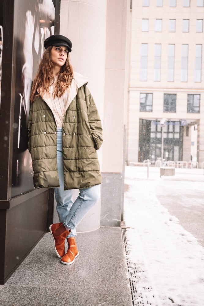 Eine Frau steht draußen im Schnee und lehnt an einem eleganten Gebäude in der Farbe QEY WEST Cognac. Sie trägt einen grünen Mantel, helle Jeans, orangefarbene Stiefel von ZAQQ und einen schwarzen Hut. Der verschneite Bürgersteig unterstreicht die frische Winterlandschaft.