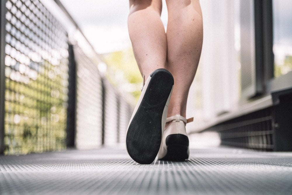 Nahaufnahme einer Person, die auf einem Metallsteg davongeht. Der Fokus liegt auf ihren Beinen und der Sohle ihres Schuhs – einem ZAQQ QITE Beige Barfußschuh aus hochwertigem Nappaleder. Der Hintergrund ist verschwommen und zeigt ein Geländer und einen Hauch von Grün.