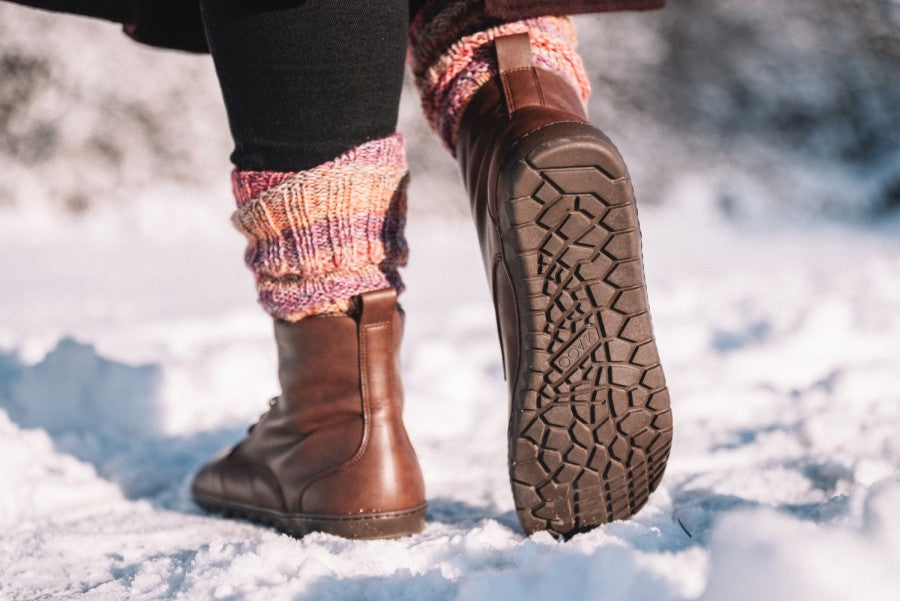 Eine Nahaufnahme zeigt eine Person, die im Schnee läuft. Sie trägt braune ZAQQ Veganer Schnürboots und gemusterte Wollstulpen. Die Sohle des rechten Stiefels, ein QUINTIC Winter Vegan Nut von ZAQQ, hinterlässt vor einem verschwommenen Schneehintergrund einen Abdruck im Schnee.