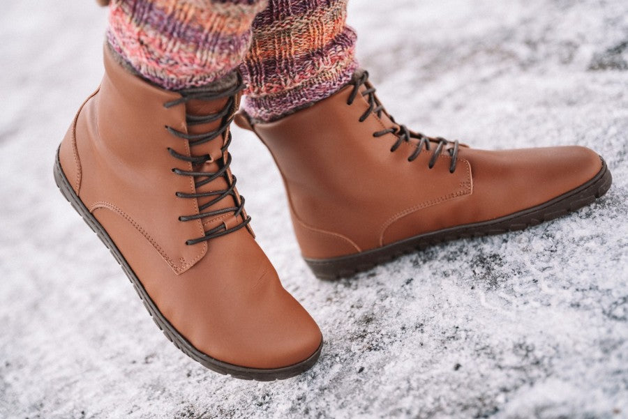 Nahaufnahme einer Person, die ZAQQ QUINTIC Winter Vegan Tabaco Stiefel mit Kautschuksohle auf einer schneebedeckten Oberfläche trägt. Die Stiefel haben eine dunkle Sohle und werden mit bunten Strickleggings kombiniert. Der Schnee bedeckt leicht den Boden und bildet einen Kontrast zum warmen Farbton des Stiefels.