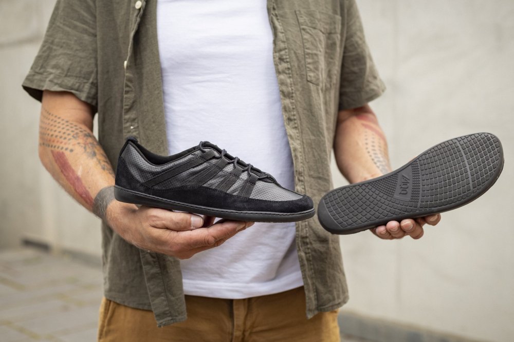 Eine Person in einem grünen Hemd und hellbraunen Hosen hält in einer Hand einen schwarzen SPARQ Low von ZAQQ und in der anderen die abgelöste Sohle. Dieser Sneaker hat ein strukturiertes Design, während die Sohle ein kompliziertes Profilmuster aufweist, und das alles vor dem Hintergrund einer schlichten Betonwand.