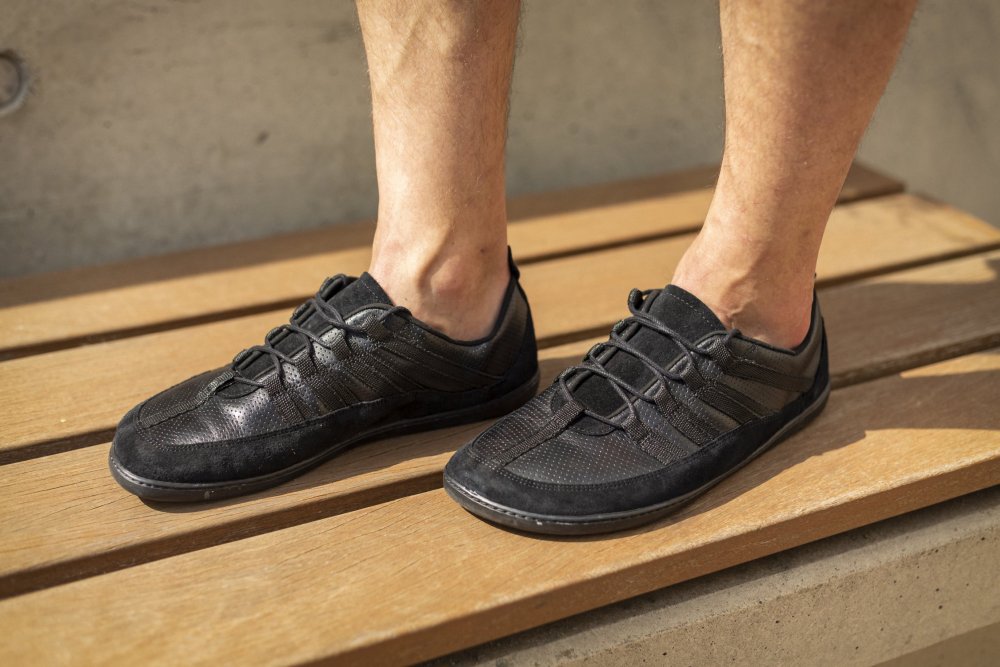 Eine Person steht auf einer Holzbank und trägt die SPARQ Low Black Sneaker von ZAQQ. Diese stylischen Sportschuhe zeichnen sich durch ein elegantes Schnürdesign mit einer Kombination aus Mesh und Leder, sichtbaren Nähten und einem minimalistischen Stil aus. Die nackten Beine der Person suggerieren eine legere Umgebung.