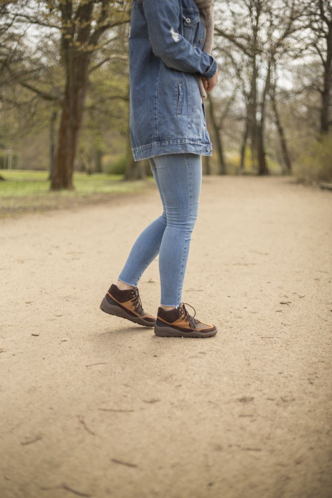 Mit den SQOUT Brown Waterproof Schuhen von ZAQQ wandert eine Person in Blue Jeans und Jeansjacke auf einem Feldweg in einem von kahlen Bäumen gesäumten Park. Der Fokus liegt auf ihrem Unterkörper und dem strapazierfähigen Schuhwerk.