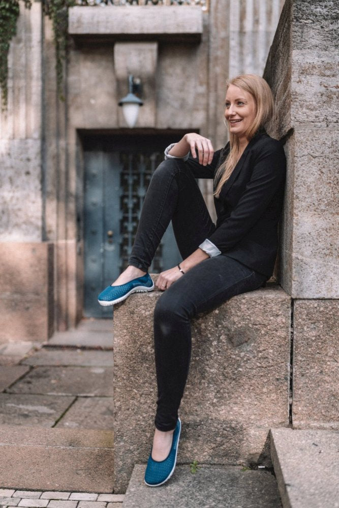 Eine Person mit langen Haaren sitzt auf einem Steinvorsprung, lehnt an einer Wand und lächelt. Sie trägt ein schwarzes Baumwoll-Strickgewebe-Outfit und dazu blaue ZAQQ SUQAR Mare-Schuhe. Im Hintergrund ist alte Steinarchitektur mit einer Tür und einer Lampe zu sehen.