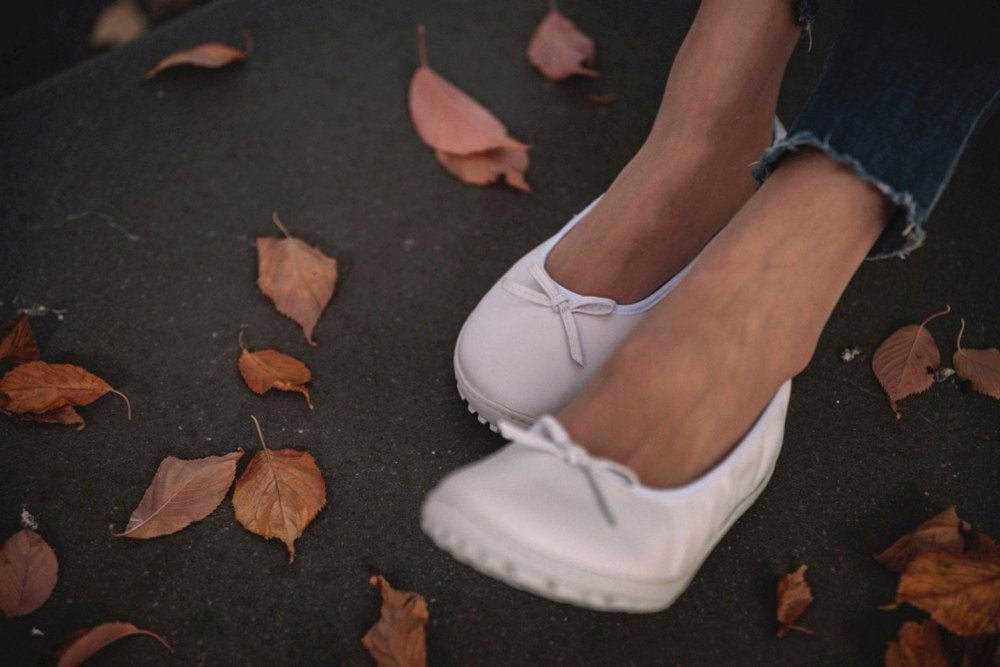 Nahaufnahme der Füße einer Person in den weißen SUQAR Flats von ZAQQ, eingebettet zwischen verstreuten trockenen Herbstblättern. Die dunklen Jeans sorgen für Kontrast, während die Schuhe mit ihrem von Nappaleder inspirierten Design die Herbststimmung verstärken.